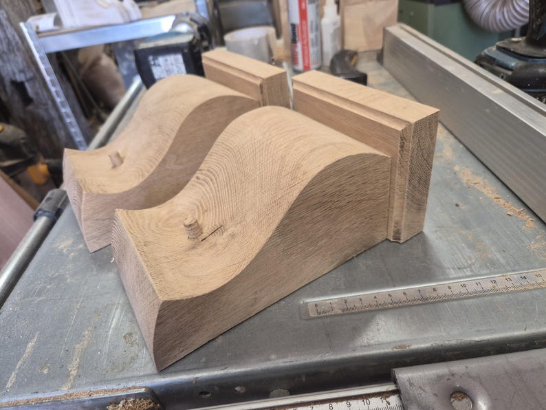 May include: Close-up of unfinished wooden furniture components. Two curved, wave-shaped pieces and two rectangular blocks are visible. The wood has a natural grain and is light brown in color. A metal ruler is in the foreground.