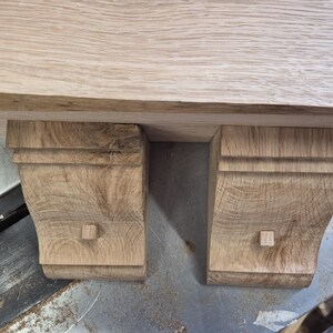 May include: A light brown oak wood shelf supported by two matching corbels. Each corbel features a curved profile with horizontal grooved detailing at the top and a small square wooden peg protruding from the center of the front face.