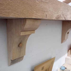 May include: A natural wood mantel shelf with two decorative corbels attached to a white wall. The corbels are made of the same wood as the shelf and are attached with wooden pegs. The shelf and corbels are unfinished.