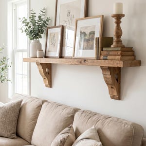 May include: A wooden wall shelf with two carved corbels holding three framed prints, a white vase with green foliage, a small jar, a wooden candle holder with a white pillar candle, and a stack of three hardcover books.