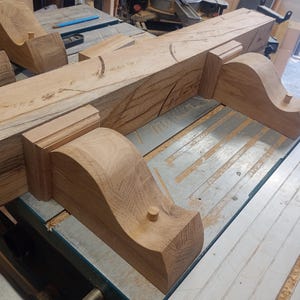 May include: Several unfinished wooden corbels and a beam are arranged on a work table. The corbels have a curved design and are made of light brown wood. The beam is also light brown and has visible wood grain. The items appear to be part of a woodworking project.