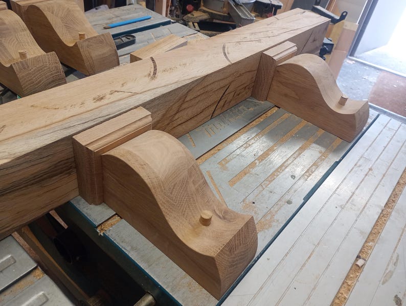 May include: Several unfinished wooden corbels and a beam are arranged on a work table. The corbels have a curved design and are made of light brown wood. The beam is also light brown and has visible wood grain. The items appear to be part of a woodworking project.