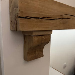 May include: A natural wood mantelpiece with a supporting bracket. The mantel is a light brown color with visible wood grain and dark cracks. The bracket is a carved, curved design, also in a light brown hue. The mantel is mounted on a white wall.