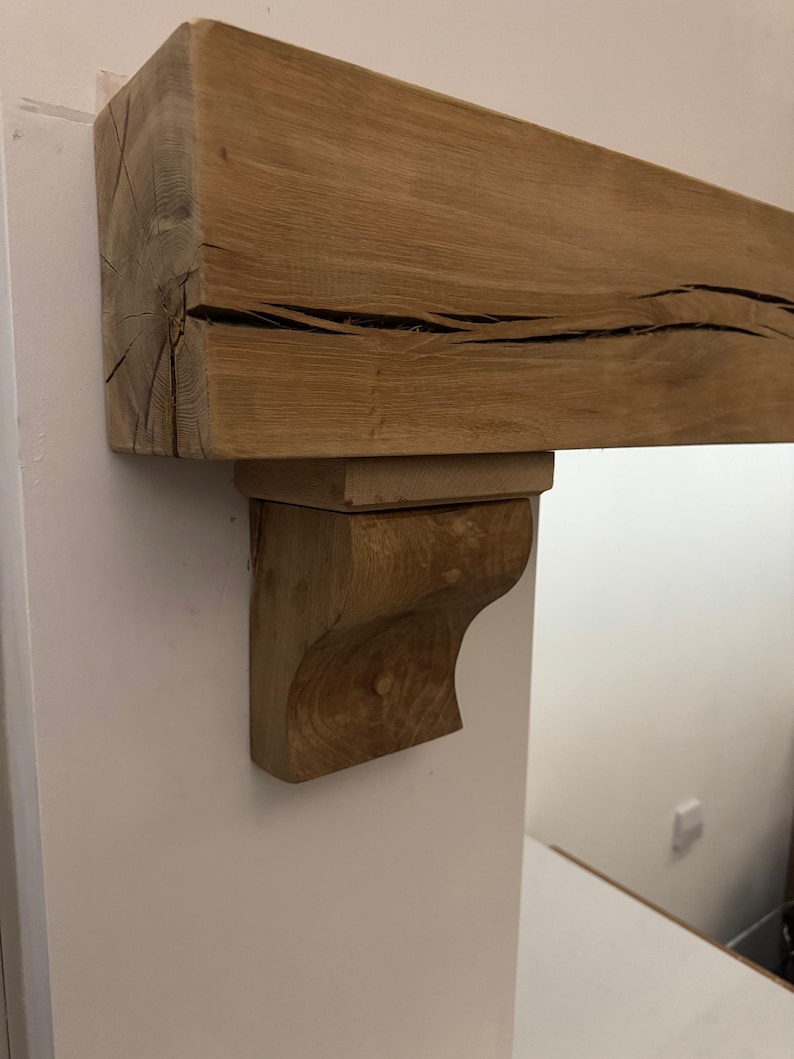 May include: A natural wood mantelpiece with a supporting bracket. The mantel is a light brown color with visible wood grain and dark cracks. The bracket is a carved, curved design, also in a light brown hue. The mantel is mounted on a white wall.