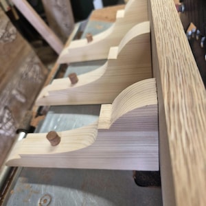 Puede incluir: Una serie de tres ménsulas de madera de color claro fijadas a un tablero vertical. Cada ménsula presenta un perfil curvo y una pequeña clavija cilíndrica de madera que sobresale de la superficie superior, mostrando las vetas naturales.