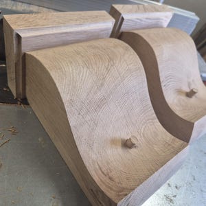 May include: Close-up of unfinished wooden corbels with a curved design. The corbels are light brown and feature a decorative, flowing shape. The wood grain is visible, and small wooden pegs are inserted into the design.