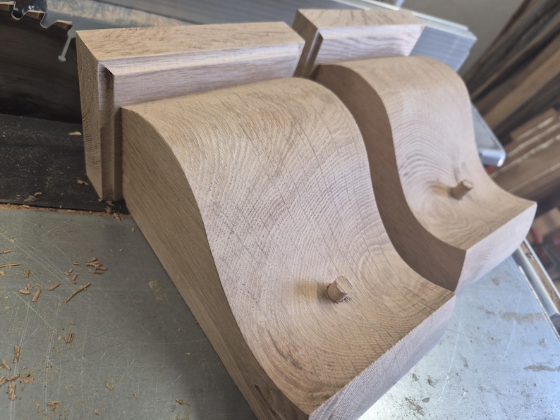 May include: Close-up of unfinished wooden corbels with a curved design. The corbels are light brown and feature a decorative, flowing shape. The wood grain is visible, and small wooden pegs are inserted into the design.