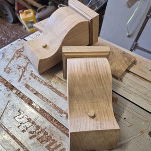 May include: Several unfinished wooden corbels with a curved design and a small wooden peg. The corbels are light brown and appear to be freshly cut. The corbels are on a work surface with sawdust.