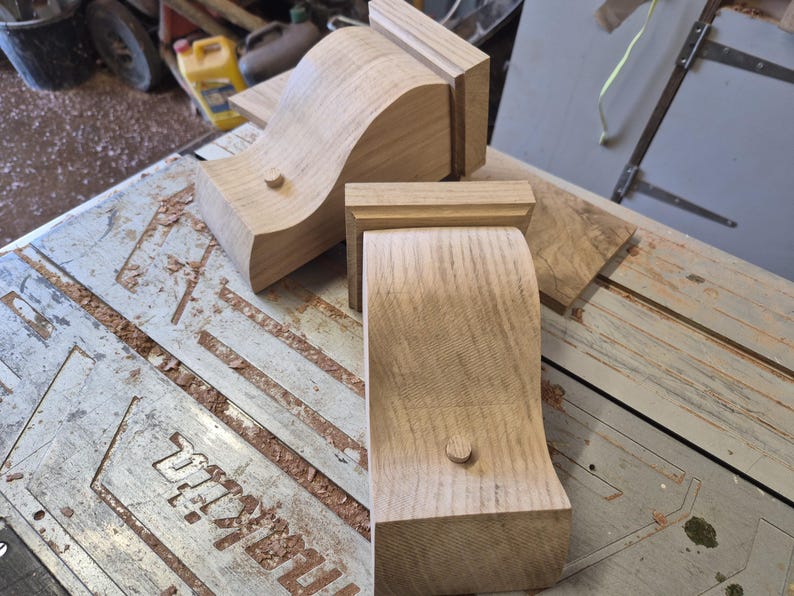 May include: Several unfinished wooden corbels with a curved design and a small wooden peg. The corbels are light brown and appear to be freshly cut. The corbels are on a work surface with sawdust.