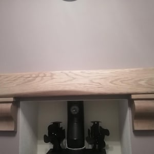 May include: A light-colored wooden mantelpiece with a curved design. The mantel is supported by two decorative brackets. Below the mantel is a fireplace opening with black decorative objects.