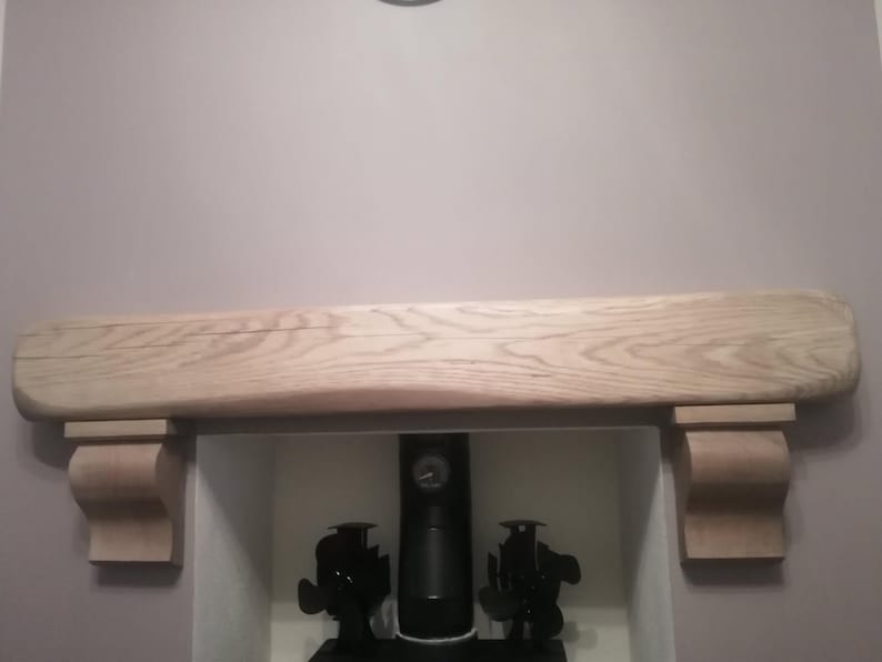 May include: A light-colored wooden mantelpiece with a curved design. The mantel is supported by two decorative brackets. Below the mantel is a fireplace opening with black decorative objects.