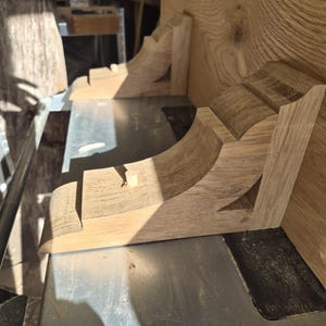 May include: Two unfinished light brown wooden corbels with a curved, stepped profile and a visible wood grain. Each corbel features a small, cylindrical wooden dowel protruding from the top flat surface, designed for structural support or mounting.