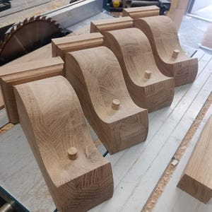 May include: Five unfinished wooden corbels with a curved design, each featuring a small wooden dowel. The corbels are light brown and sit on a light-colored surface, alongside a piece of wood and a circular saw blade.