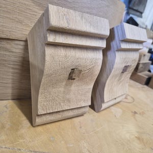 Könnte beinhalten: Zwei unfertige Holz-Konsolen mit dekorativem Design. Die Konsolen sind hellbraun und haben eine geschwungene Form mit einem quadratischen Detail. Sie liegen auf einer hellbraunen Oberfläche.