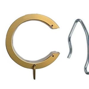 May include: A gold-colored circular curtain ring with a small attached loop at the bottom and a separate silver-colored metal pin hook with a sharp pointed end, designed for hanging curtains on a rod with a matching track system.
