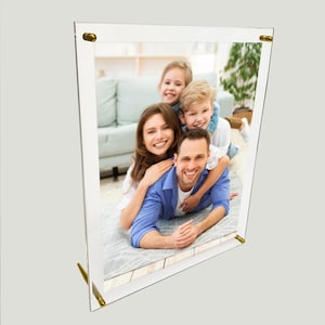 May include: A clear acrylic photo frame with gold accents holds a photo of a family of four. The family is smiling and appears to be having fun together. The frame is standing upright on a white surface.