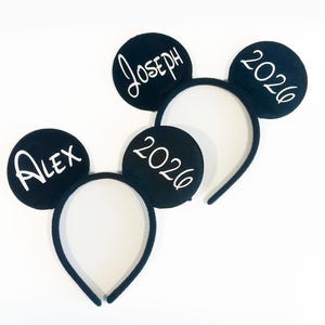 May include: Two black headbands with circular ears. One headband features the name ALEX and the year 2026 in white lettering. The second headband features the name JOSEPH and the year 2026 in white lettering. The headbands have a textured fabric finish.