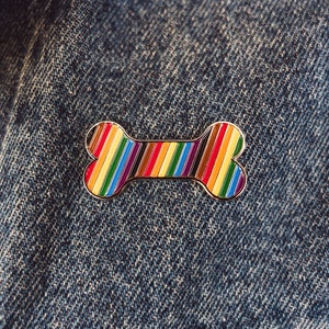 May include: A rainbow striped dog bone pin on a blue denim background.