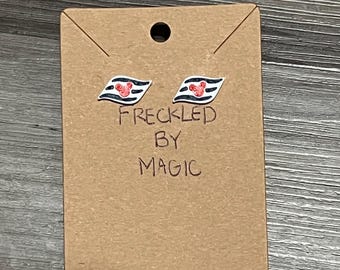 May include: A pair of stud earrings featuring a red heart with Mickey Mouse ears on a black and white striped background. The earrings are displayed on a brown card with the text "Freckled By Magic".