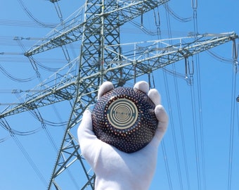May include: A hand wearing a white glove holds a black and gold sphere with a swirling pattern. The sphere is in front of a large metal electrical tower against a blue sky.