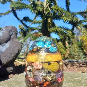 Puede incluir: Una pieza de arte de resina transparente en forma de huevo, llena de capas de piedras de colores, cristales y copos de oro. La capa superior presenta piedras azules, mientras que el centro tiene una banda dorada con un símbolo geométrico dorado.