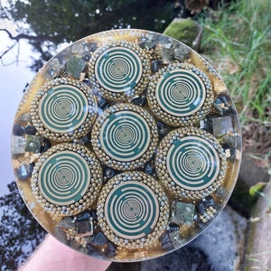 Könnte beinhalten: Eine kreisförmige Harzscheibe mit sieben eingebetteten grünen und goldenen kreisförmigen Leiterplattenmustern. Jedes Muster ist von kleinen Metallperlen umgeben. Die klare Harzbasis ist mit dunkelgrauen Steinen und Goldflocken gefüllt.