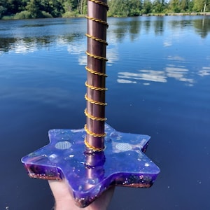 May include: A decorative star-shaped object with a purple base and a copper-coloured pole wrapped with gold wire. The object is held in front of a lake with trees and a blue sky in the background.