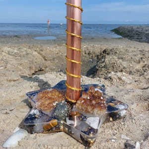 Puede incluir: Un tubo de cobre envuelto en una bobina de alambre dorado, colocado verticalmente sobre una base de resina en forma de estrella. La base contiene racimos de cristales color ámbar, piedras metálicas oscuras y pequeñas cuentas incrustadas.