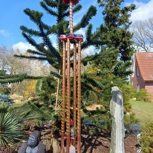 Op de afbeelding: Een hoge, koperkleurige buitensculptuur met een rode en zwarte boven- en onderkant. De sculptuur heeft een cilindrisch ontwerp met een spiraalvormig draadpatroon en is gevuld met kristallen en stenen. De sculptuur is omgeven door groen en andere tuinornamenten.
