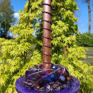 Puede incluir: Una pieza de arte artesanal de resina púrpura con una columna central de color cobre envuelta con alambre púrpura. La base es una plataforma circular púrpura llena de piedras y cristales coloridos. El fondo presenta follaje verde y un cielo azul.