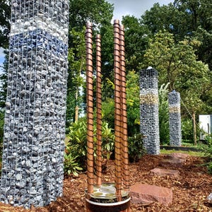 Op de afbeelding: Een tuinbeeld met vier verticale koperen buizen omwikkeld met draad, geplaatst op een cilindrische voet met horizontale banden van koper en zilverkleurig metaal. Op de achtergrond drie hoge rechthoekige schanskorven gevuld met grijze en witte stenen.