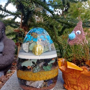 Puede incluir: Un generador de energía orgónica en forma de huevo con capas de piedras, cristales y detalles dorados. La parte superior presenta motas turquesas y doradas. Un símbolo dorado de la flor de la vida está centrado en la parte delantera. La base es de color marrón claro.