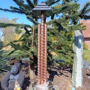 Op de afbeelding: Een koperen windgong met een piramidevormige top en spiraalvormige accenten. De windgong staat in een glazen kom met water en kristallen. De achtergrond omvat een gorillastandbeeld, een stenen sculptuur en een groene boom.