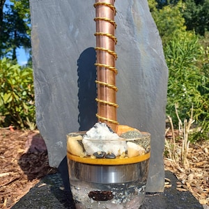 Puede incluir: Un tubo de cobre cilíndrico envuelto en una bobina de alambre dorado, colocado verticalmente dentro de un recipiente de resina transparente. El recipiente contiene piedras blancas, beige y marrones, escamas metálicas y una banda amarilla.