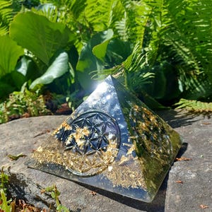 Peut inclure: Une pyramide en résine translucide dotée d'une base noire avec des flocons de feuilles d'or incrustés et un symbole circulaire de fleur de vie sur la face avant. L'objet présente une surface lisse et polie qui reflète la lumière.