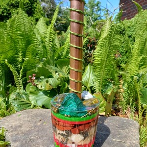 May include: A decorative item with a clear resin base, layered with red, brown, and green sections. A copper rod wrapped in green wire rises from the base. The item is displayed outdoors, surrounded by green plants.