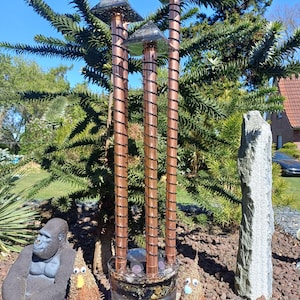 Op de afbeelding: Een tuindecoratie met drie koperkleurige solar lichten met piramidevormige toppen. De lichten staan in een transparante glazen container met decoratieve elementen. Een gorillastandbeeld en draadfiguren zijn ook aanwezig.