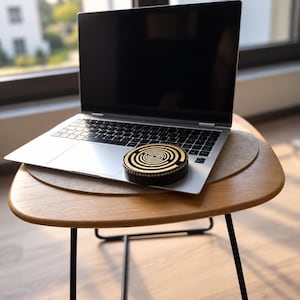 Może przedstawiać: Srebrny laptop z czarnym ekranem stoi na drewnianym stole. Na klawiaturze laptopa znajduje się okrągły, czarno-złoty przedmiot dekoracyjny z wzorem koncentrycznego labiryntu na górnej powierzchni oraz kropkowaną krawędzią.