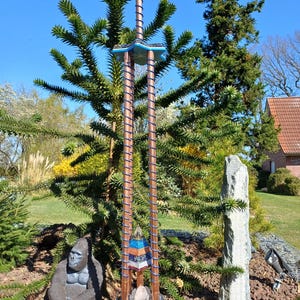 Peut inclure: Une sculpture décorative haute en cuivre et en verre. La sculpture présente une série de formes géométriques empilées, y compris des pyramides et des cubes, et est ornée de cristaux et de pierres colorés. La sculpture est placée sur un fond d'arbres verts et de ciel bleu.