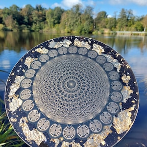 May include: A round, decorative plate with a dark blue background and gold leaf accents. The plate features a complex geometric design in the center and around the edges. The plate is held up in front of a lake and trees.