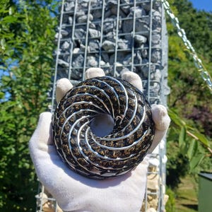 Può includere: Un oggetto circolare a forma di toro con una superficie scura e strutturata con pagliuzze color oro incastonate e un intarsio continuo a spirale in filo metallico color argento che avvolge l'intera forma, creando un motivo geometrico.