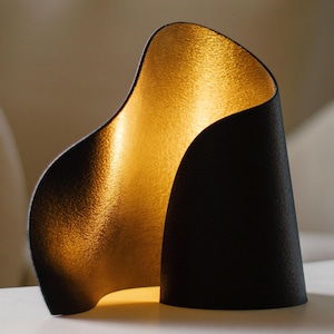 May include: A modern black table lamp with a gold interior. The lamp has a curved, sculptural design, with a textured exterior and a warm, golden glow emanating from within. The lamp is on a white surface.