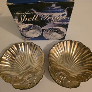 May include: Two silver-plated shell trays with a matching box. The trays are in the shape of a scallop shell with a detailed design. The box is dark blue with the words "Old Hampshire Silversmiths" and "Silverplated Shell Trays".