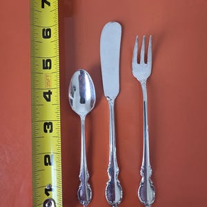 May include: A silver spoon, butter knife, and fork set with ornate handles. The spoon measures approximately 4 inches, the knife is 6 inches, and the fork is 6 inches. The silverware is displayed on a reddish-brown surface.
