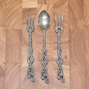 May include: A set of antique silver-colored flatware, including two forks and a spoon, arranged on a wooden surface. The utensils feature ornate, detailed handles with floral and scrollwork designs. The spoon has a rounded bowl, and the forks have three tines.