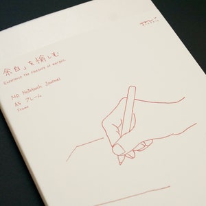 Puede incluir: Una cubierta de cuaderno blanca con texto rojo que dice "『余白』を愉しむ Experience the pleasure of margins. MD Notebook Journal A5 フレーム Frame". Un dibujo de línea roja de una mano sosteniendo un lápiz está en la cubierta.