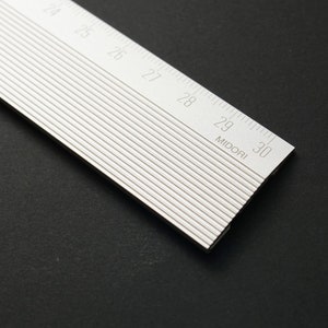 Könnte beinhalten: Ein silbernes Lineal aus Metall mit schwarzen Markierungen. Das Lineal ist 76,2 cm lang und hat Markierungen für jeden Zentimeter. Das Lineal ist mit "MIDORI" an der 73,66 cm-Marke gekennzeichnet.