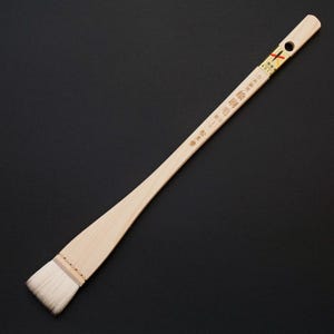 Puede incluir: Un pincel de caligrafía largo, con un cabezal ancho y rectangular y un mango delgado. El mango es de madera clara y tiene texto japonés y un pequeño agujero. El pincel tiene cerdas blancas.