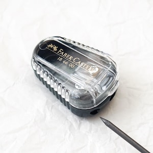 May include: A clear plastic pencil sharpener with a black base. The sharpener has a silver trim and the text "FABER-CASTELL 18 66 00" printed on the side.
