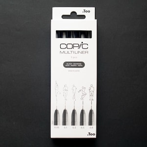 May include: A white box of Copic Multiliner pens, with the brand name and product details printed on the front. The box contains five black pens with different tip sizes, ranging from 0.05 to 0.8. The box also features illustrations of figures.